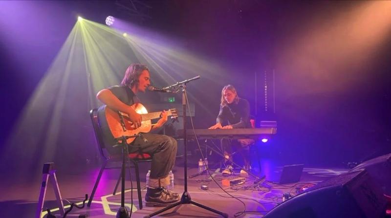 On stage under lights, keys beside an acoustic guitarist